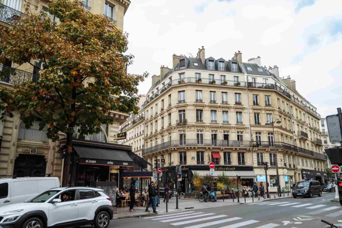 Une rue animée de Paris avec un immeuble haussmannien, un restaurant à l'angle et des piétons, idéale pour une pause avant de rejoindre l’Aéroport Roissy CDG.