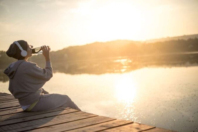 Une personne avec un casque audio est assise sur un ponton en bois et prend le temps de rester hydraté face au lac au coucher du soleil.